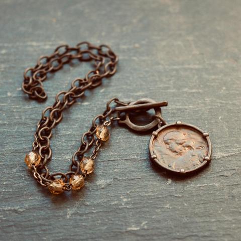 A photo of a blackened pewter pendant necklace on a dark grey slate background. The pendant features a weathered image of Joan of Arc; the thick linked chain contains a fragment of 4 amber coloured glass rosary beads and a rustic toggle closure.
