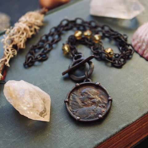 A photo of a blackened pewter pendant necklace lying on a vintage textured hardcover book and is surrounded by raw crystal points and shells. The pendant features a weathered image of Joan of Arc; the thick linked chain contains a fragment of 4 amber coloured glass rosary beads and a rustic toggle closure.