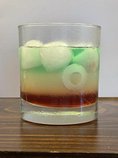 A rocks glass on a dark wooden surface against a white backdrop. It's mostly full of a liquid and some ice. The drink is in three roughly equal horizontal layers. The bottom layer is a dark red, the middle is off-white, the top is a bluish green.