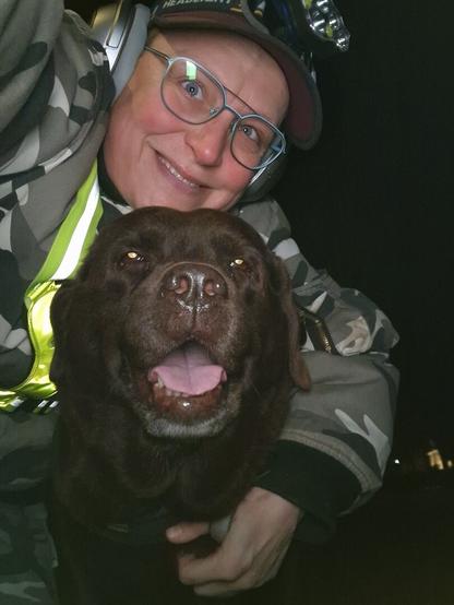Night time selfie of a person and a chocolate brown Labrador with greying snout. The person is wearing a white-grey military camouflage jacket, yellow safety harness, red cap, silver headphones and blue-rimmed glasses. On their cap rests a headstrap torch in off position. Their free hand is around the dog's shoulders. Both are facing the camera. The dog's mouth is slightly open, showing off some mlem. The person is smiling, they're grateful for the time spent together with their lovely gall.