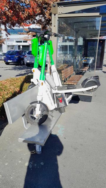 Three Lime branded (hire-by-app) electric scooters stacked on top of each other at various angles atop a bus-stop seating area, locked in place by the uppermost having it's front wheel wedged behind the seat-back.