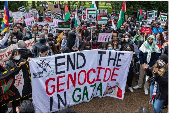 A crowd of pro-Palestine protesters holding pro-Palestine signs with a giant flag with the words "END THE GENOCIDE IN GAZA" on it.