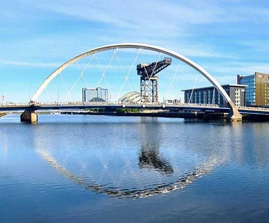 A curved bridge against a blue sky reflected in calm waters of the Clyde in Glasgow.