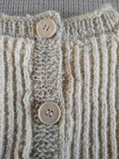 Des boutons gris clairs sur une bandes de boutonnage d'un gris un peu plus foncé. Le reste du tricot est fait de bandes blanches et grises.