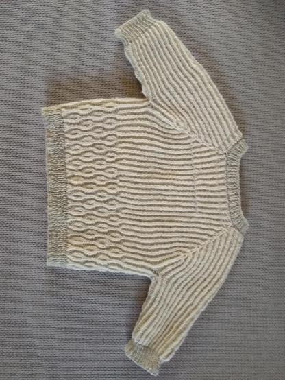 Le dos d'un gilet à manches longues pour bébé. Le col est gris clair. L'empiècement et le corps sont en mailles blanches et grises. Les mailles blanches forment des colonnes du haut de l'empiècement jusqu'au milieu du dos puis se croisent pour former des bulles.