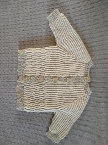 L'avant d'un gilet à manches longues pour bébé. Le col et les pattes de boutonnages sont grises. Les mailles sur l'empiècement, le corps et les manches sont blanches et grises. Elles forment des colonnes de couleurs. Au milieu du devant les colonnes se croisent pour former des bulles.