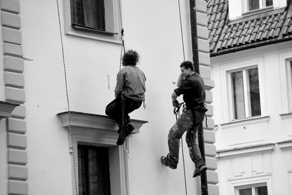 Dans une rue dont les maisons ont des façades blanches. Deux hommes accrochés à des filins, sont suspendus au niveau du premier étage L'un est au dessus de la porte d'entrée moulurée et un peu en dessous de la fenêtre qui la surplombe..  A un mètre l'un de  l'autre, au milieu de cette ascension,  ils semblent poursuivre leur conversation en toute tranquillité.