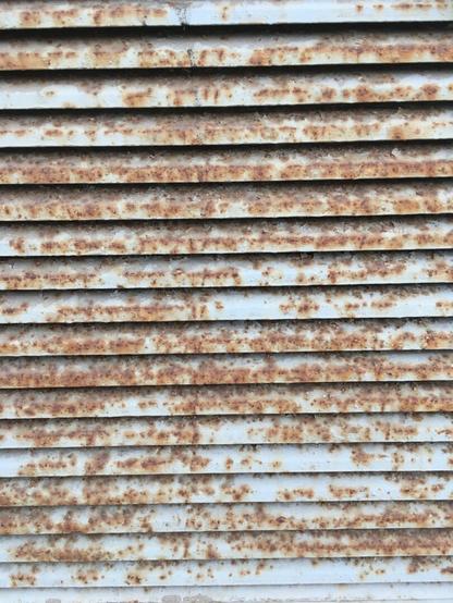 A close-up image of a rusted metal louvered vent or grille, showing multiple horizontal slats with significant corrosion and reddish-brown rust patches.
Deutsch: Nahaufnahme eines verrosteten Lamellengitters aus Metall mit mehreren horizontalen Lamellen, die starke Korrosion und rostbraune Flecken aufweisen.