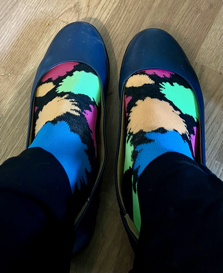 Photo of 2 feet wearing dark blue slip-on shoes and black socks with cheerful splotches of green, orange, pink, and blue at the end of black pants.