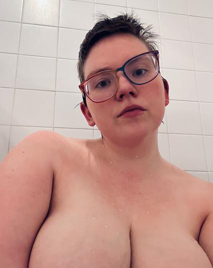 A naked nonbinary person with short hair and crooked rainbow glasses is seen in front of a white tiled wall, from the mid-chest up. They have extremely large breasts, seen from just above the areola, and their skin appears slightly wet.