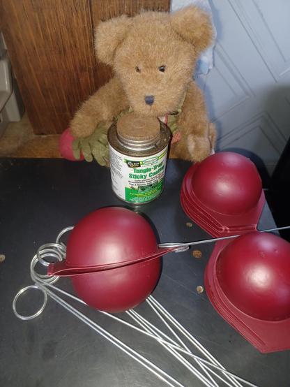 A ten inch tall (25cm), light brown teddy bear in green overalls sits looking directly at the camera. His name is BeetBear. He is holding a beet in his right paw.  There is a bunch of beets embroidered on the bib of his overalls.  In front of him is a can of "Tanglefoot Sticky Stuff"  There are red plastic shells that can be folded so the two halves make a sphere that can be fitted in to the wire holders laying in front.