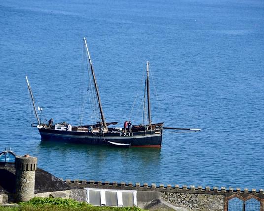 Three masted wooden sailing boat painted mostly black with a white stripe just below the gunwales seen side on with sails down. It’s moored  just off shore in calm blue waters in front of a historic house with crenelated garden walls and the black roof and circular distinctive chimney on the left. 

Two crew are busy working at the front if the boat. The registration  PZ.17 in hand drawn white lettering can be read on the side of the boat towards the front.