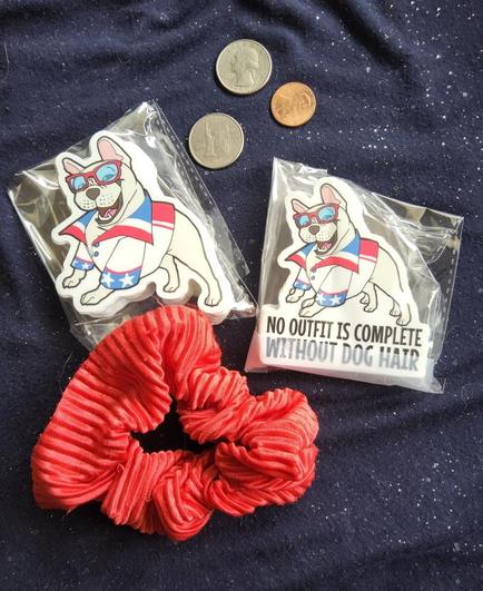 Flat lay photo of a french bulldog dressed in a red white and blue Super Dave-esque outfit. On a sparkly blue fabric, with some assorted coins for scale and a red scrunchie.