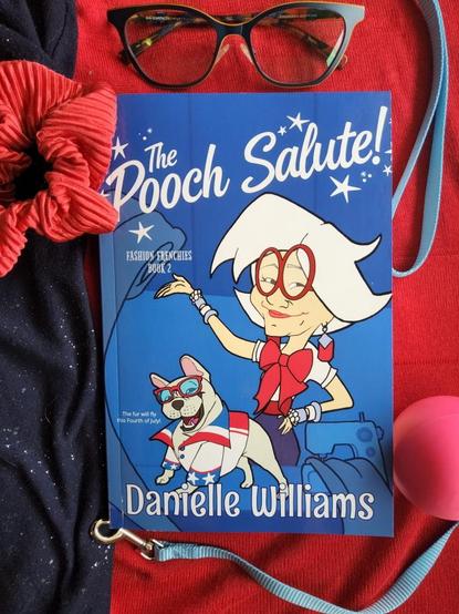 Flat lay of my novel "The Pooch Salute!" against red and sparkly blue fabric. At the top, there's a pair of glasses, to one side, a blue leash and pink rubber ball, and to the other side, a red scrunchie.