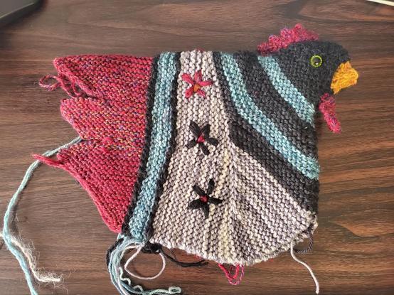 A red, gray, black and light blue knitted chicken laying flat, prior to stuffing