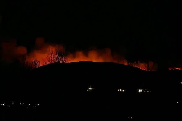 A wide shot of a dark landscape at night with the bright orange glow of a wildfire illuminating the horizon behind a dark silhouette of a hill. Some scattered lights are visible in the foreground.
