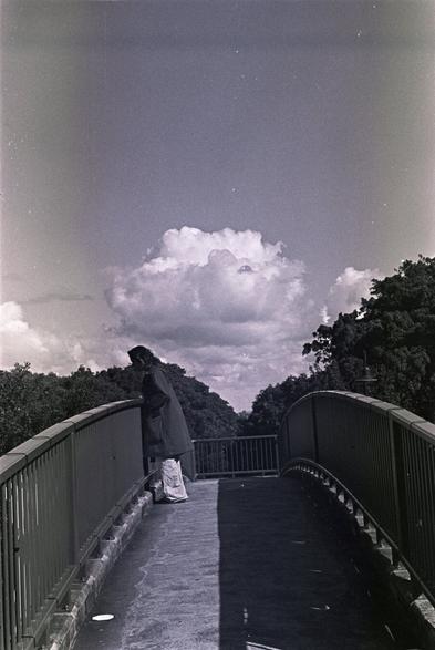 A woman in a coat looks down over a pedestrian overpass, tall winter skies behind her. B&W photo.