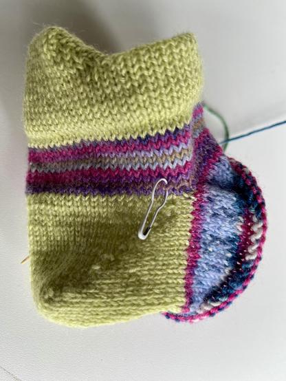 Sock in the making. It has a folded heel tab and short row heel in bright green and a few rows on the leg and foot in a variegated yarn with purples, pinks and blues. There's a bulb stitch marker close to the corner of the heel.