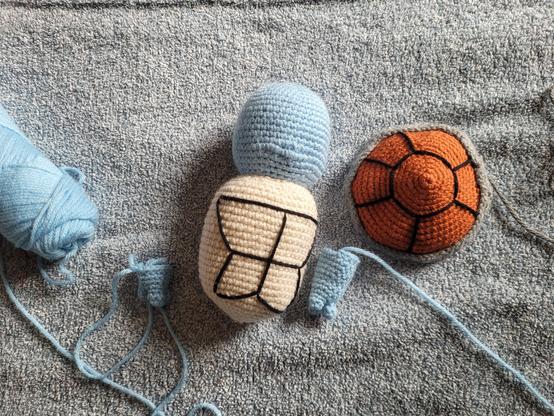 A light tan shell with black lines embroidered on it along with a light blue head without details sewn on.  The back of the shell in a brownish-orange with a grey edge is next to it also embroidered with black yarn for the segments.  Next to the body is one complete arm and one in progress arm.