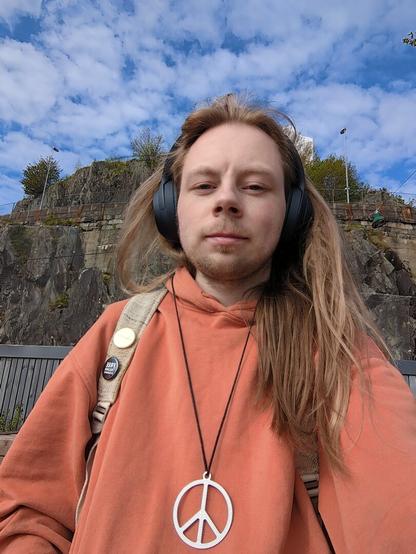 A person wearing headphones and an orange hoodie, a peace symbol pendant, and headphones outdoors against a backdrop of a rocky hillside under a partly cloudy sky.

The scene is a selfie-style portrait, with the person positioned in the center of the frame.

The person is looking directly at the camera, with their expression being neutral.
