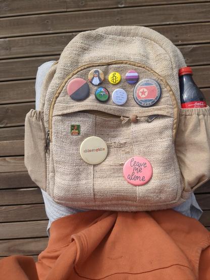 The beig backpack is positioned centrally in the image, with various pins of different shapes and colors attached to its surface.  

The backpack's texture appears rough and natural, and it's situated against a backdrop of light brown wooden planks.

The pins on the backpack depict diverse symbols, including political and social awareness. Some are themed, while others are more abstract. The colors of the pins vary, creating a vibrant contrast against the backpack's neutral tone.