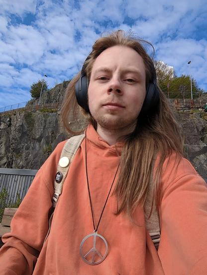 A person wearing headphones and an orange hoodie, a peace symbol pendant, and headphones outdoors against a backdrop of a rocky hillside under a partly cloudy sky.

The scene is a selfie-style portrait, with the person positioned in the center of the frame.

The person is looking directly at the camera, with their expression being neutral.