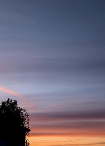 A serene sunset scene fills the frame. The lower portion of the image glows with a vibrant orange hue, transitioning upwards into soft pink and then into a deep, dusky blue. Horizontal bands of cloud stretch across the sky, adding texture and depth to the gradient of colour. In the bottom left corner, the silhouette of a tree and the edge of a building add a touch of contrast against the bright sky. The overall impression is one of tranquility and natural beauty.