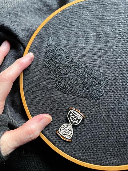 Lia’s fingers show a 2.5 by 3.5 inch wonky blob of black on black whipped backstitch that looks a bit like flames, but if you’ve been following this piece you’ll know it’s pubic hair. It’s definitely pubic hair. An hourglass-shaped needle minder that reads “this is taking forever” is attached to the cloth.