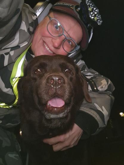 Night time selfie of a person and a chocolate brown Labrador with greying snout. The person is wearing a white-grey military camouflage jacket, yellow safety harness, red cap, silver headphones and blue-rimmed glasses. On their cap rests a headstrap torch in off position. Their free hand is around the dog's shoulders. Both are facing the camera. The dog's mouth is slightly open, showing off some mlem. The person's head is resting on the dog's head. They're smiling, grateful for their time spent together.