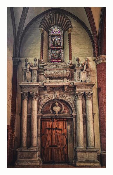 A wooden door surrounded by ornately carved stone statues in a church. In the centre bottom of the picture there is a wooden door with black metal hardware. On either side of the door there are two stone carved columns. On top of the door there is a carved stone bust to a male religious figure. Above this again there are more statues and carvings of angels and a coffin-like object. Above the carvings and stonework on the wall is a brightly coloured stained glass window depicting Jesus and the Franciscan monks.