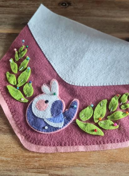 A work in progress of a pink banner flag, a simplistic felt cockatiel design pinned on the point at the bottom and green leaves surrounding it.