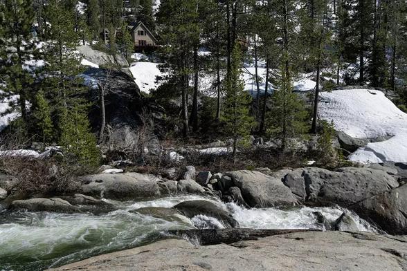 Photograph of a small river rushing through rocky, high-altitude terrain. A copse of evergreen trees and a slope covered with snow are visible in the background. Photo credit: Penny Collins for the Associated Press.