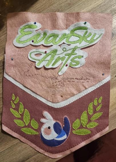 Picture of an unfinished felt hanging banner flag. 
It is mainly dark pink and a soft pink, a simplistic cockatiel is sewn onto the bottom, green leaves on each sides of it. 
On the top part, the words « Evan Sky Arts » are machine embroidered; under it, another line of text is embroidered in dotted lines.
