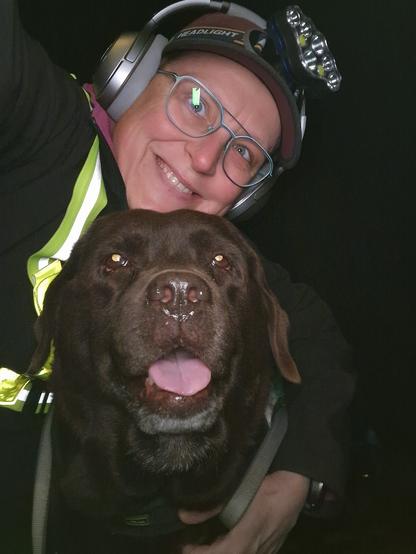 Night time selfie of a person and a chocolate brown Labrador with greying snout. The person is wearing a black jacket, yellow safety harness, red cap, silver headphones and blue-rimmed glasses. On their cap rests a headstrap torch in off position. Their free hand is around the dog's shoulders. Both are facing the camera. The dog's mouth is slightly open, showing off some blep. The person's head is resting on the dog's head. They're smiling, grateful for their time spent together.