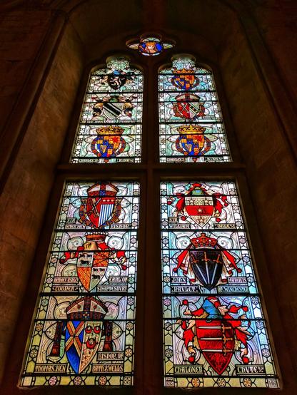 A stained glass window in the Great Hall, Winchester. The window is tall and narrow, with multiple panes. Each pane contains a different heraldic shield, with colourful designs and inscriptions. The shields are arranged in a grid pattern. The background is a dark, stone wall.