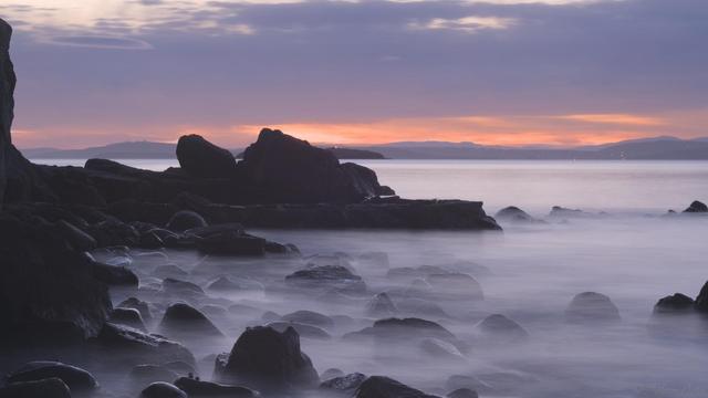 View across a bouldered shoreline at sunset, a long exposure turning the waves to a wispy smoke, drifting around the wet, glistening rocks in the foreground.  A grey blue cloud above turning orange at the horizon across the bay.