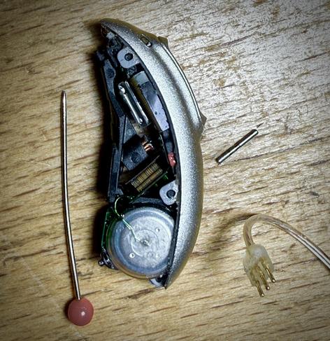 side view of hearing aid with bottom plastic cover removed. There’s round button batteries, maybe a coil or two, some kind of electronics “sandwich” circuit boards, and small round catches holding the top on. A push pin is shown for scale and the wire with three leads/pins leading to actual ear speaker is on bottom right