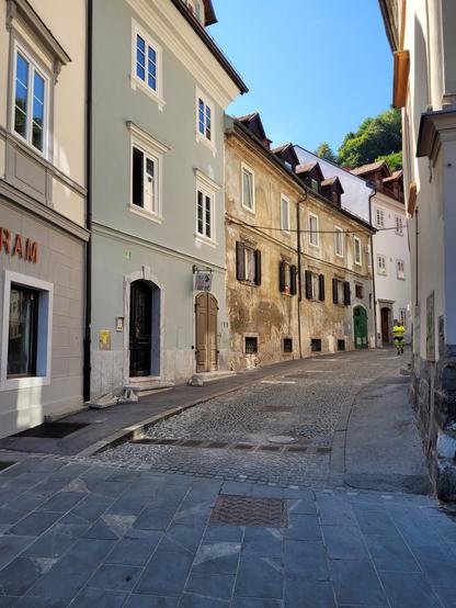 Hochformat in Farbe. Blick in eine, nach rechts geschwungene, Altstadtgasse. Links und rechts stehen alte Wohnhäuser mit 3 bis 4 Geschossen. Die Häuserfassaden auf der rechten Seite sind durch die Biegung kaum zu sehen. Auf der linken Seite stehen als erstes zwei Häuser mit neu gemalter Fassade,das erste bräunlich, das zweite Grün. Das Haus danach ist stark verwittert, das letzte Haus frisch weiß gestrichen. Die Häuser stehen lückenlos nebeneinander und sind unterschiedlich hoch. Die Straße zwischen den Häusern besteht aus einem mittleren Streifen mit Pflastersteinen und zwei schmalen Asphaltstreifen vor den Häusern. Der Bodenbelag vorne im Bild besteht aus rechteckigen Platten. 
Hinten in der Gasse tritt ein Mann um die Biegung. Er trägt kurze, neongelbe Arbeitskleidung und schaut während des Gehens auf seine Armbanduhr.
Oben zwischen den Häusern ist strahlend blauer Himmel zu sehen, sowie die Kronen einiger sommergrüner Laubbäume.