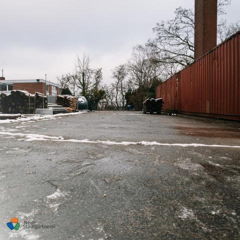 Ein teilweise vereister Asphaltboden mit vereinzelten Schneeresten. Rechts  rote Container und ein Ziegel-Schornstein, der in den Himmelragt. Links ein flaches Gebäude, davor Stapel mit Holz/Baumstämmen. Im Hintergrund kahle Bäume und ein grauer Winterhimmel.