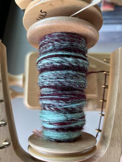 Light wooden bobbin on a spinning wheel with a slubby single on it made of a burgundy longwool mixed with a bright mint downy short staple wool.