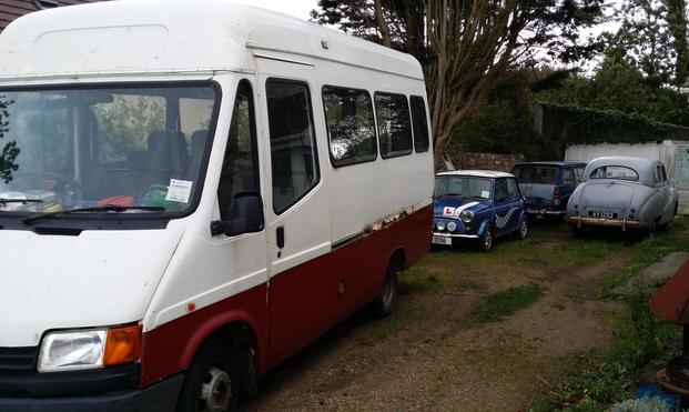 Four elderly motor vehicles in grassy driveway with trees in the background. Nearest to the camera is a Transit Minibus with a total of five side windows including the door. The top half is white, the bottom colour I'd describe as rust, were it not for the layer of rust that separates the two.

Behind it a tidy blue BMC Mini with a white roof and wide wheel arches is in front of a dark blue Morris Oxford Estate. The tailgate is a paler blue or grey, probably primer, and the rear fins and nicely chromed tail lights are all in order. 

To the right of the Mini and Oxford sits an Austin Somerset, grey and curvacious but fairly solid, although the rear bumper is rusting nicely.