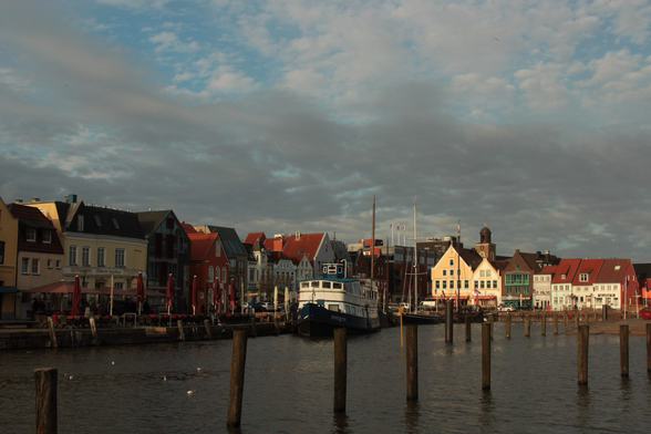 Farbfotografie im Querformat zeigt einen Blick in den Husumer Hafen. An der Mole sind mehrere unterschiedliche Schiffe festgemacht (Fähren, Segelschiffe, Motorboote) vor den bunten Häusern, die den Hafen umgeben. Der Himmel ist hellblau und bewölkt.