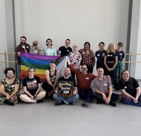 2 rows of people pose with a progress pride flag and a painted sign for the Queer Tango Collective. 9 stand in the back, and 7 sit in front.