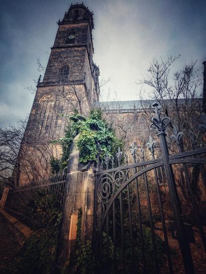 A low-angle shot captures a tall, gothic-style church under a dramatic, cloudy sky. The church, constructed from dark stone, features a prominent tower with a clock face. The base of the church is partially obscured by a decorative wrought-iron fence and some greenery, including ivy climbing the stone walls. The overall tone is moody and atmospheric.