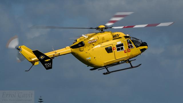 Yellow rescue helicopter manoeuvring in sunlight from right rear. It is banked to the right and angled up, giving a top view of the white and red striped rotors.