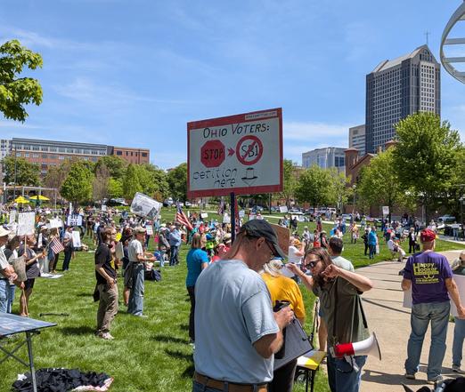 A crowd of people in a park for a protest and rally, with a man in the foreground with a sign saying,"Ohio voters, stop SB1.!"