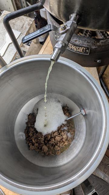 Wort slowly streams into a conical fermentor, foaming up where it's landing. Crushed wine pomace can be seen in the bottom of the fermentor.