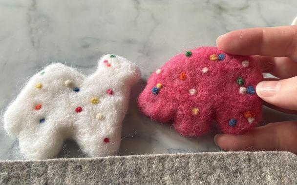 two needle-felted replicas of Mother’s Circus Animals cookies: a white camel and a pink bear, both with rainbow sprinkles. they’re about 2.5 times the size of actual cookies.
