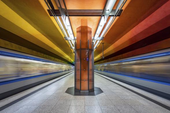 Symmetrical shot of two blue metro trains crossing each other on each side of an empty plaftorm painted in yellow and orange at Candidatz metro station in Munich, Germany.  A big orange pillar separates the two trains. The shot was taken with a wide angle lens to strech the perspective in the corner of the images and with a slow shutter speed to capture the movement of the train and give an impression of high speed.