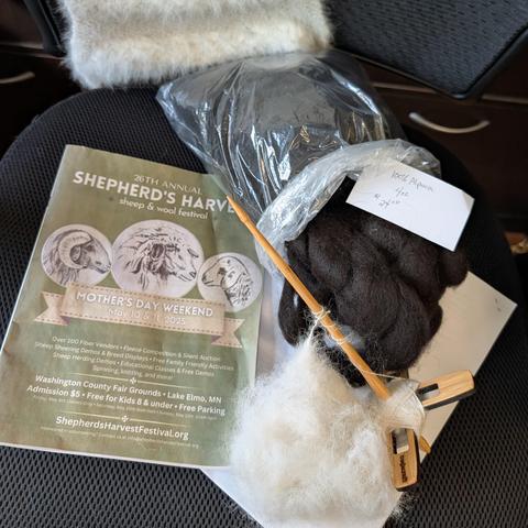 Ribbony blobs of shiny black fiber emerge from a plastic bag. A tag reads 100% Alpaca, 4 oz, $24.00. It sits next to a Turkish spindle holding a fine white single spun from a cloud of husky fiber. There's also a brochure for the Shepherd's Harvest Sheep and Wool festival, and a bit of a white husky yarn scarf in the background.